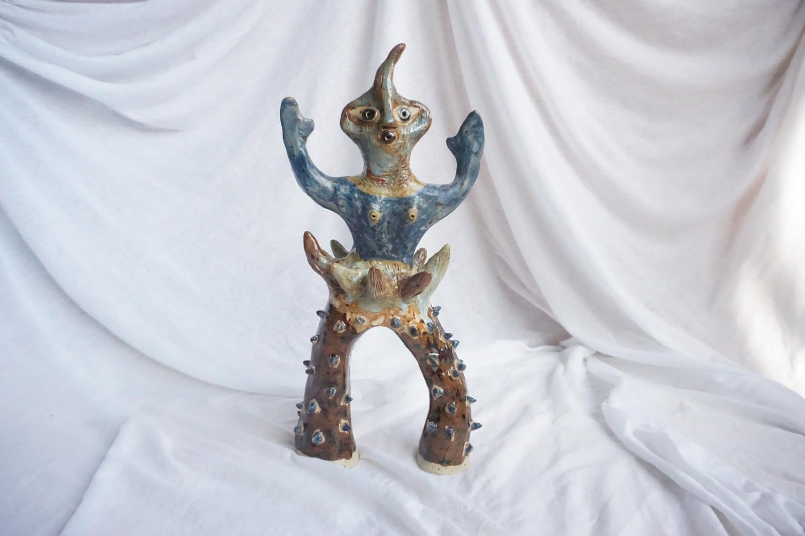 Glazed ceramic creature by Livia De Magistris, arms raised, standing on two spiked, arched legs.